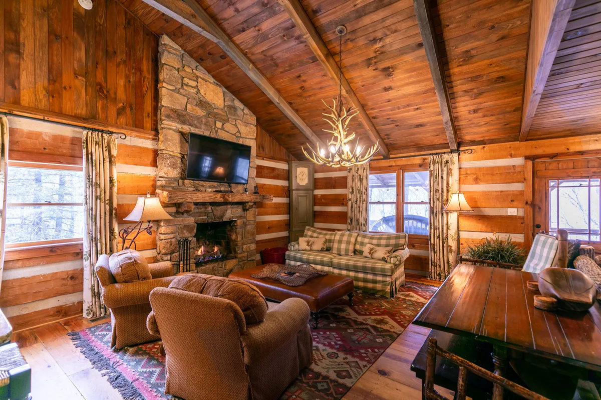 A warm, inviting great room showcasing a stone fireplace, antler chandelier, plaid sofa, and a large wooden dining table. The area is staged for relaxing or entertaining, with ample seating for groups and visible décor touches that evoke a mountain retreat vibe.
