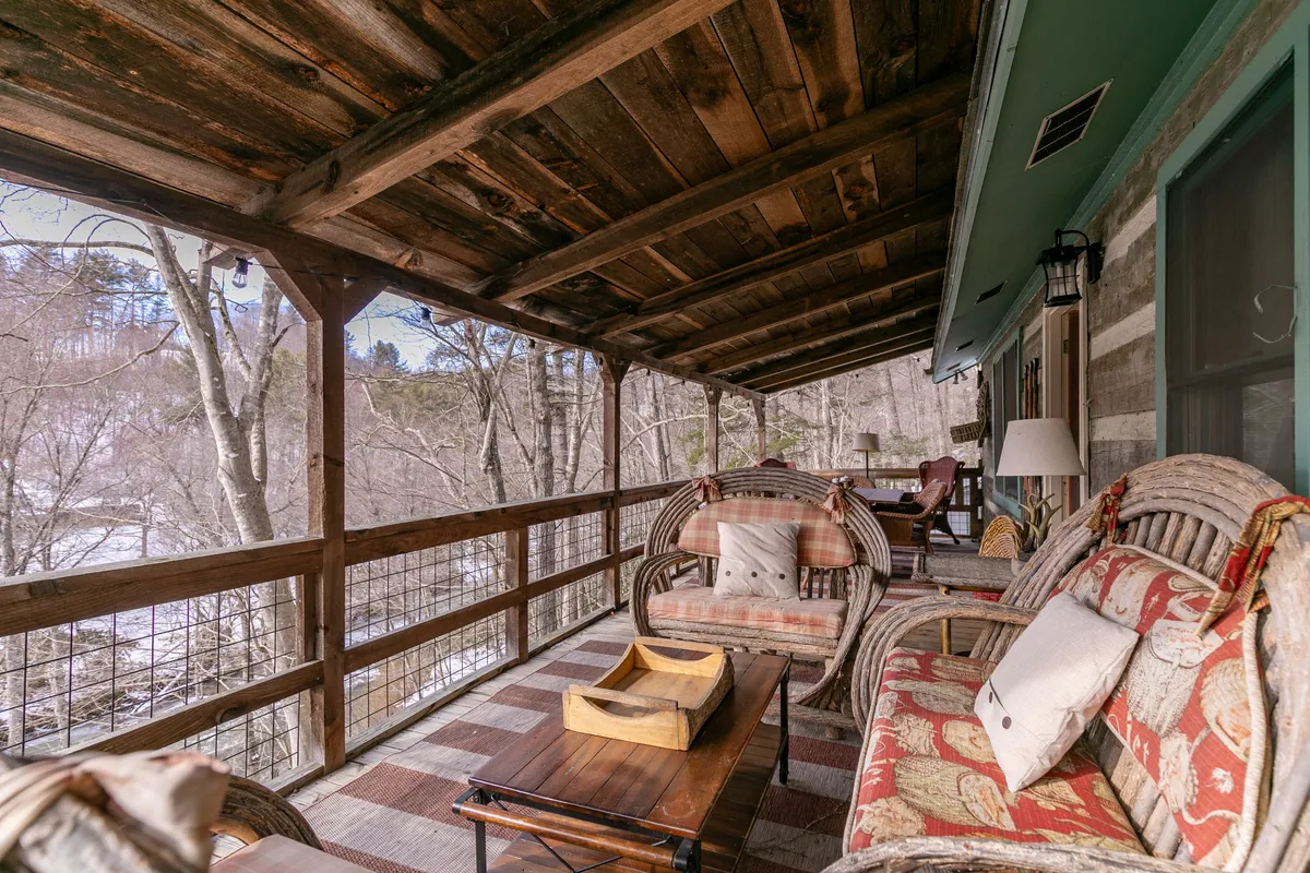 This image features a cozy deck space with comfortable seating, decorative cushions, a coffee table, and rustic wooden beams. The area is surrounded by treetops, creating a peaceful, elevated retreat. The scene invites guests to gather, relax, enjoy sunsets, morning coffee, or host intimate conversations in a natural setting.