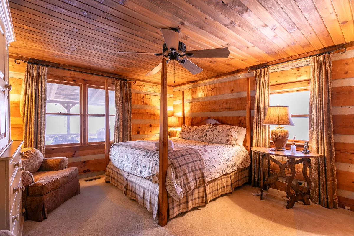 A warmly lit bedroom featuring a four-poster bed with plush linens, a wooden ceiling, table lamp, and large windows. The cozy setup highlights a space designed for relaxation and quiet evenings, with natural light streaming in.