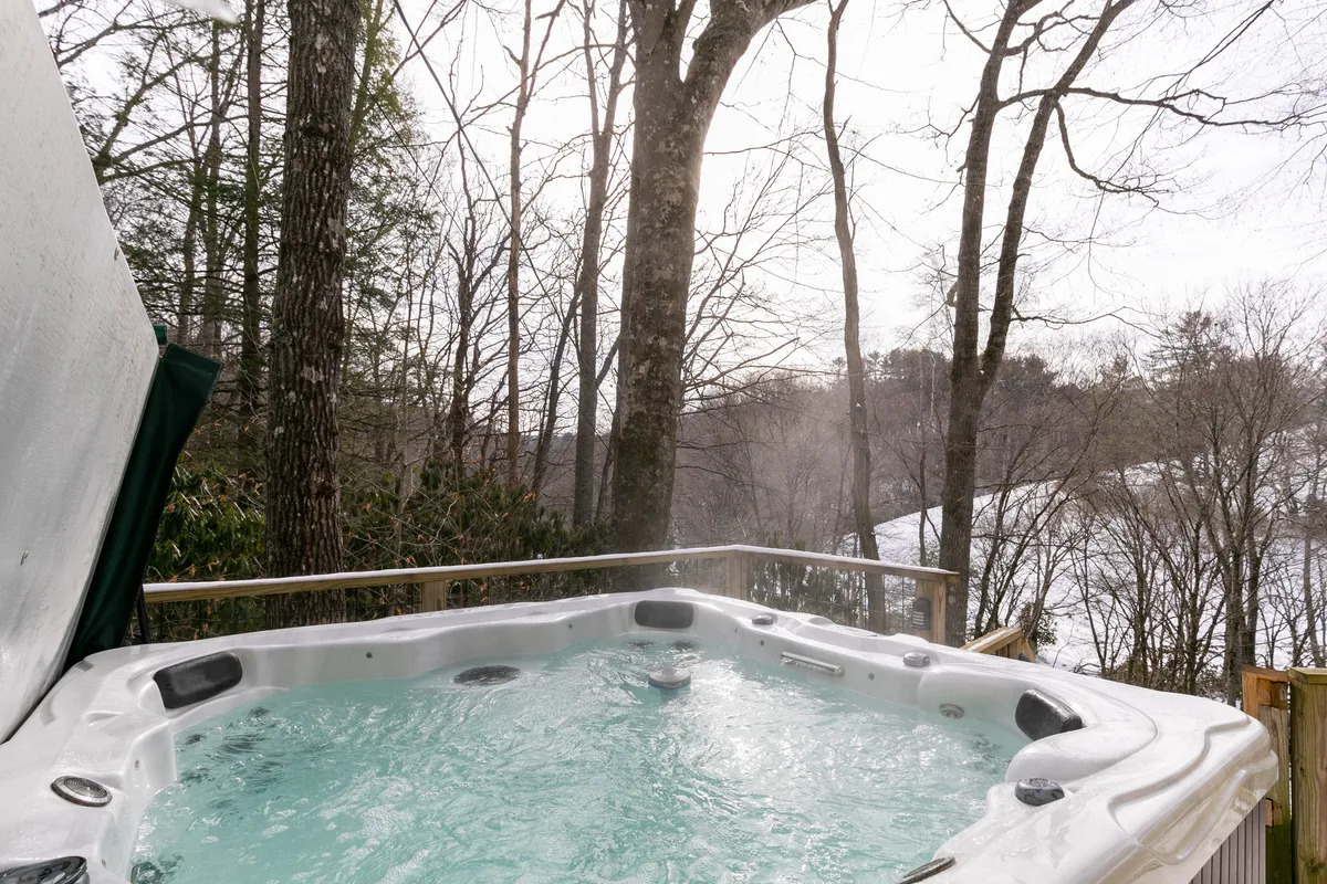 The image showcases an outdoor hot tub surrounded by trees and distant hills, with steam rising and serene mountain views beyond. The setting is tranquil, offering an inviting spot to unwind after a day of adventure or skiing.