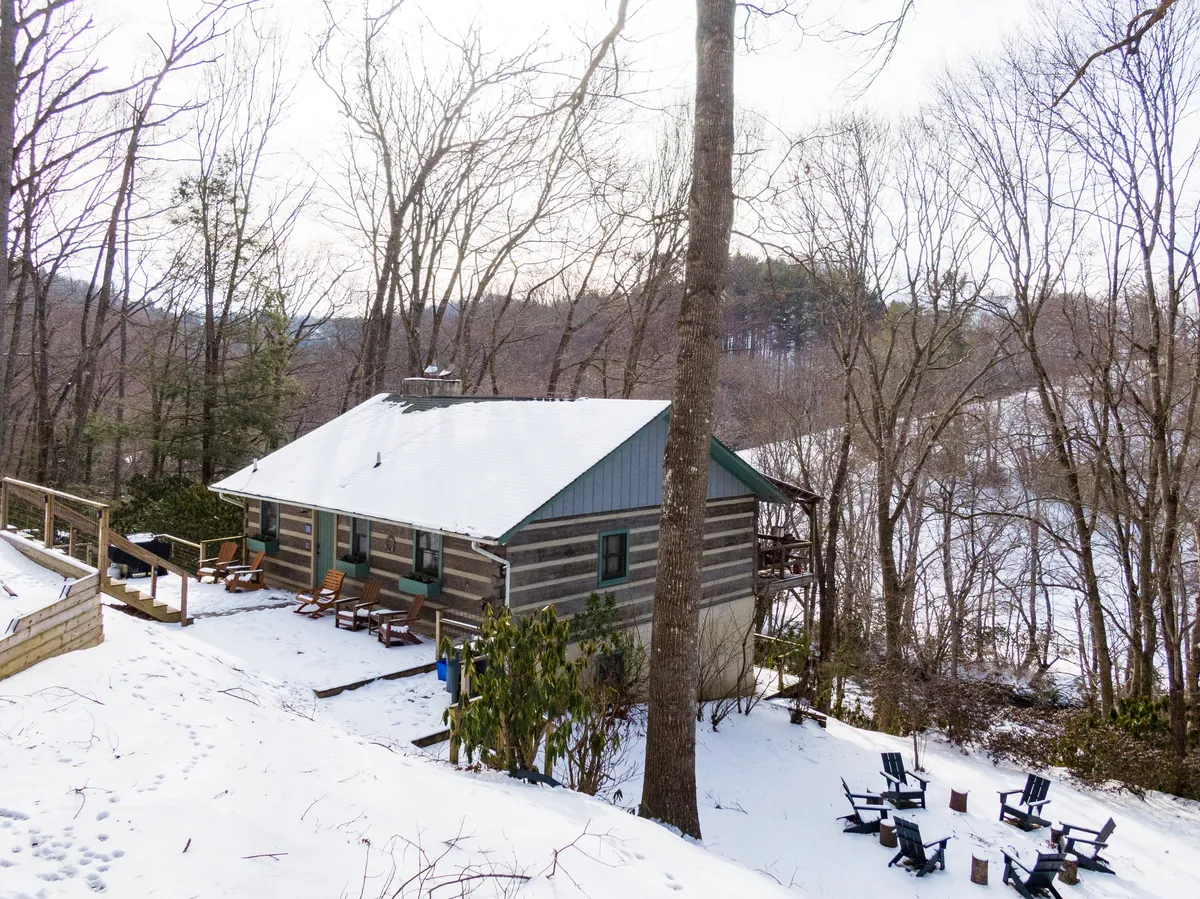This exterior image features a charming log cabin with a wooden deck, outdoor seating, and a snow-covered ground, evoking the cozy and serene vibe of a winter mountain retreat. The deck is perfect for relaxing by the fire pit or sipping hot cocoa while watching snowfall.