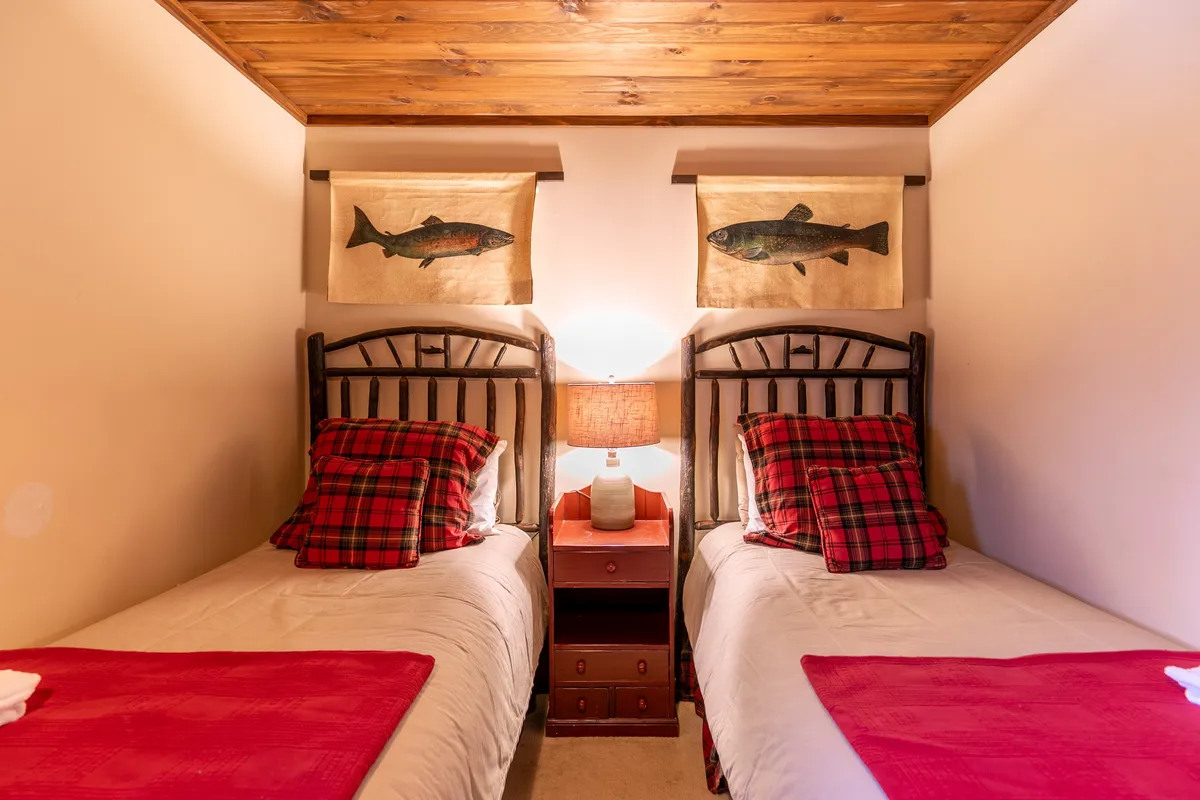 A welcoming bedroom with twin beds, adorned with plaid bedding and fish-themed wall art. The room features a shared lamp and a soft, inviting ambiance—ideal for children, friends, or flexible sleeping arrangements.