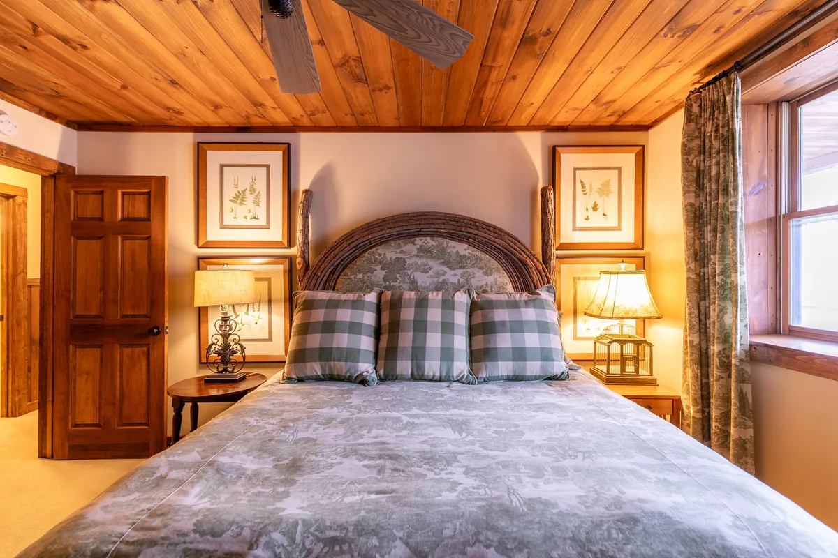 A restful bedroom scene with a comfortable bed adorned with decorative pillows, a wooden ceiling, framed artwork, and soft lamp lighting. The space feels private and is perfect for reading or quietly unwinding.