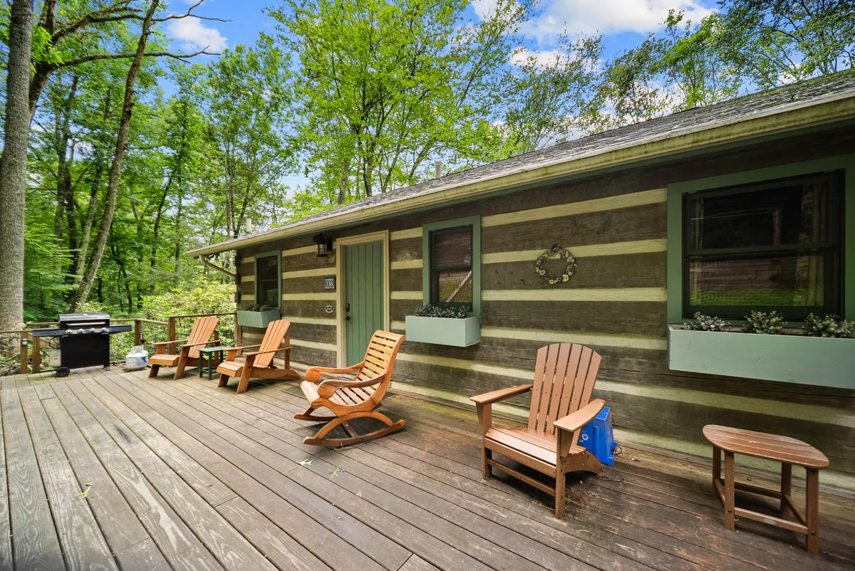 The image showcases the rustic log cabin exterior nestled amidst lush green foliage. A spacious wooden deck with inviting Adirondack chairs overlooks the natural beauty, offering a serene setting for guests to relax, sip coffee, or gather for sunset barbecues by the river.