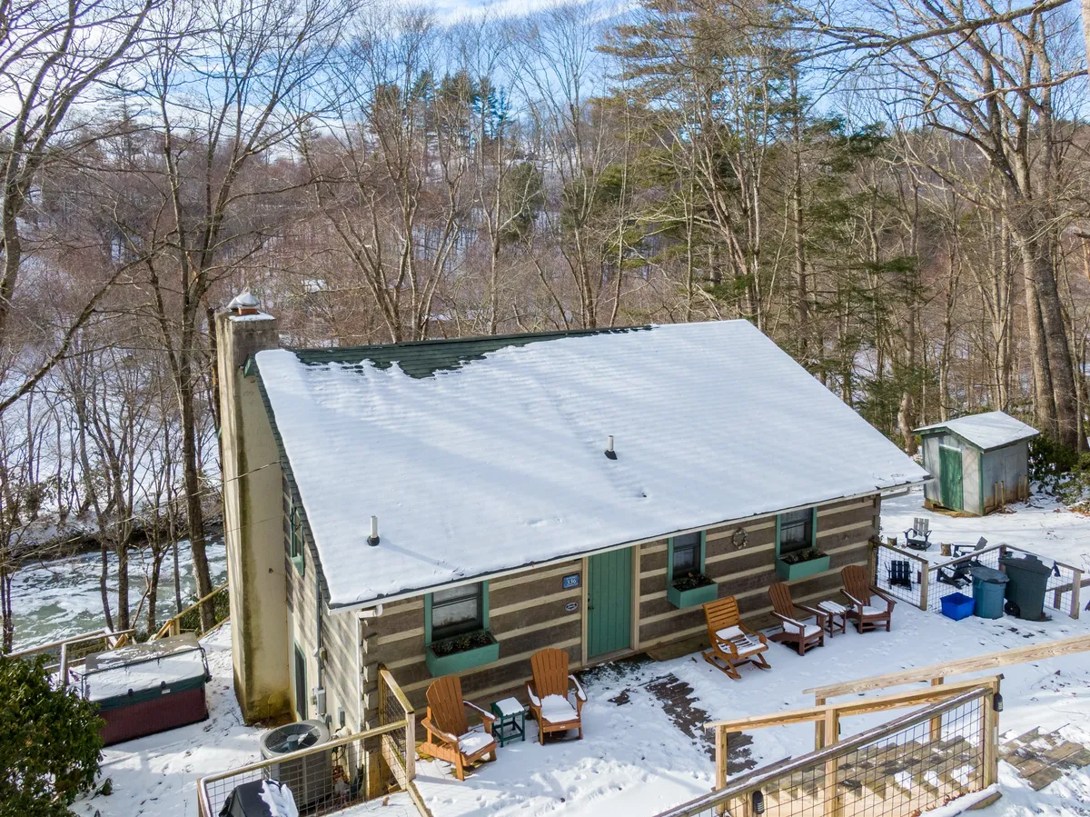 A cabin structure with a snow-covered roof and wooden chairs set on a porch, surrounded by wintertime trees. The scene is tranquil, highlighting both the comfort of the cabin and its snowy, secluded location.