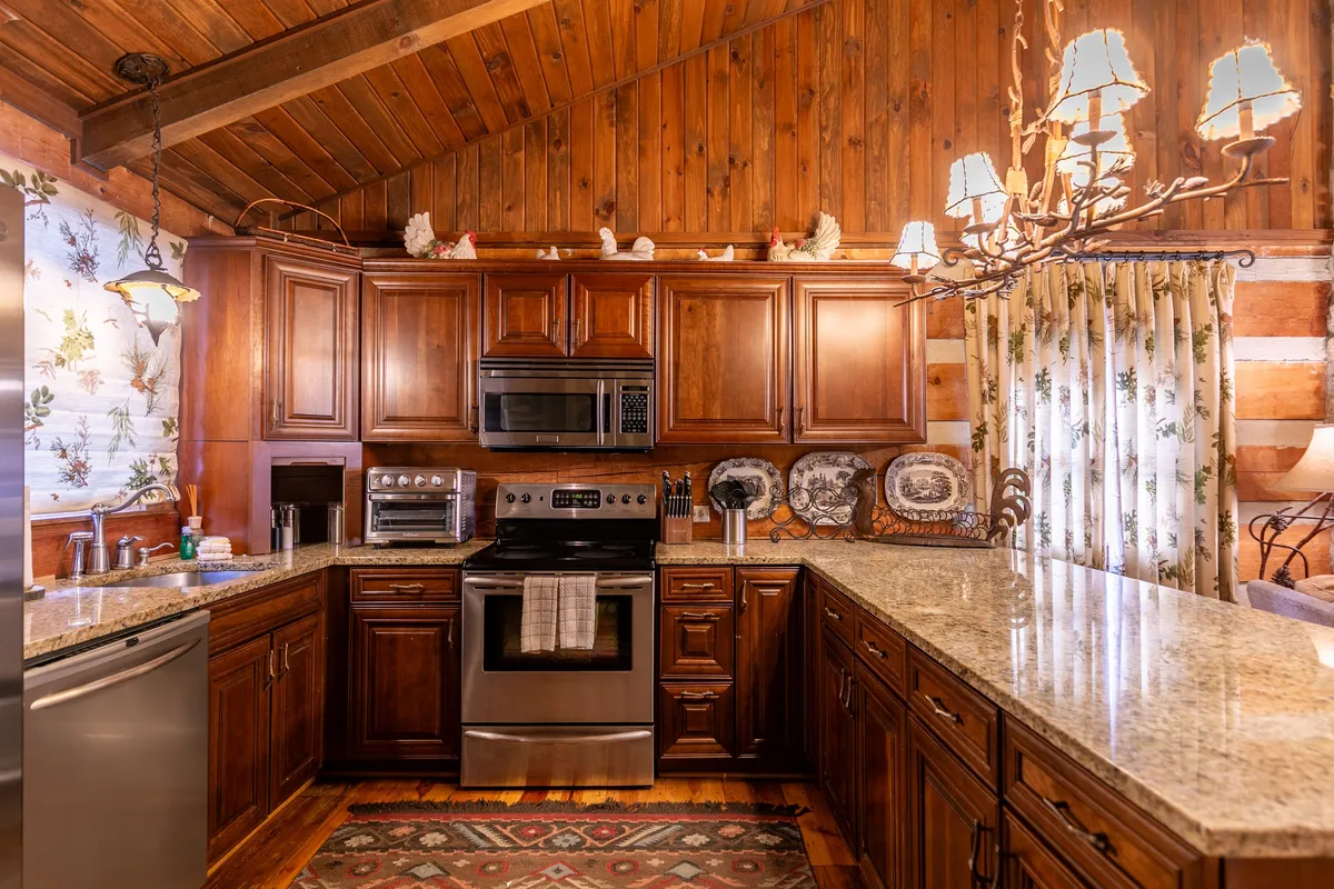 A warm, cozy kitchen with custom wooden cabinetry, granite countertops, and elegant lighting fixtures. Natural light streams through a window framed by floral curtains, illuminating the inviting workspace. The kitchen exudes a homey atmosphere, perfect for cooking and gathering.