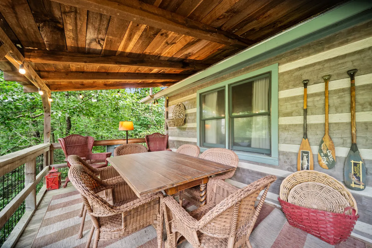 A wooden outdoor dining table surrounded by woven chairs sits on a covered deck, accented by decorative paddles on the wall. Lush greenery provides a tranquil backdrop, and the scene feels welcoming and ideal for group gatherings or quiet moments. The space exudes comfort and a strong connection to the natural surroundings, matching the description of the main level's outdoor living and dining area overlooking the river.