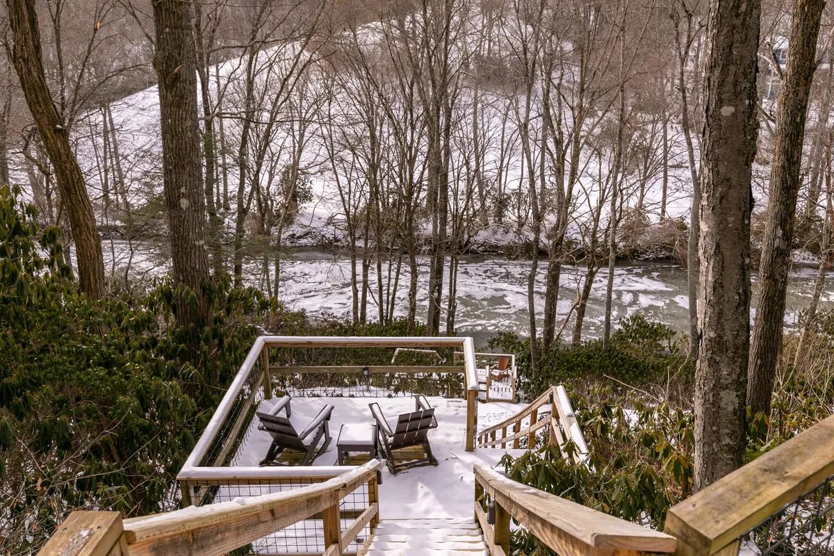 A wooden deck with chairs overlooks a snowy river and bare trees, creating a peaceful spot to relax, observe wildlife, or enjoy the tranquil river views just steps from the home.